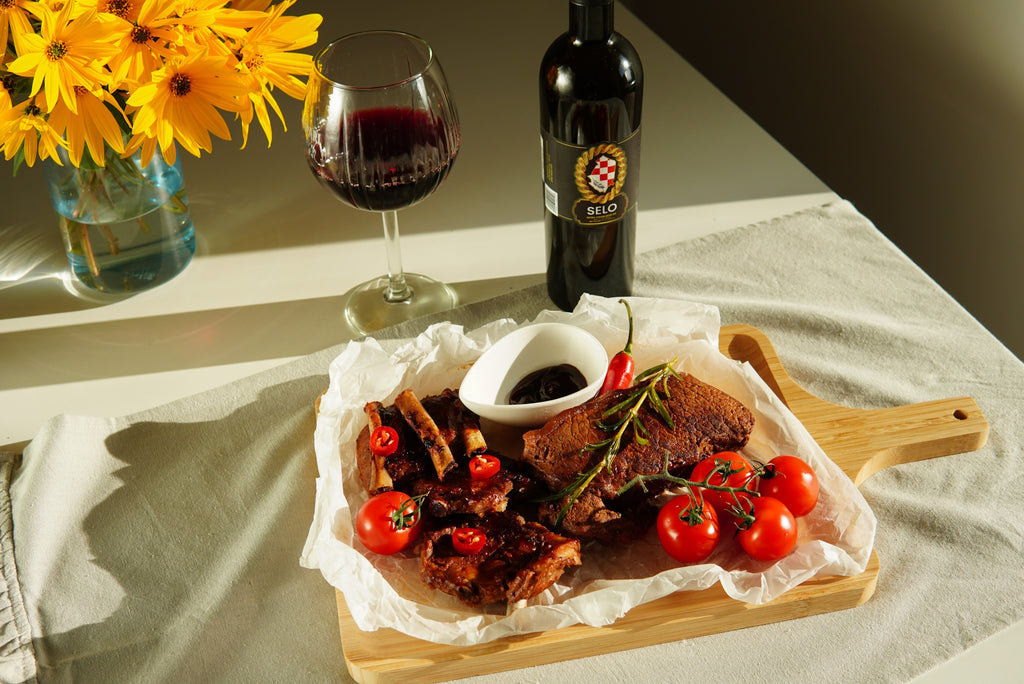 A plate of grilled meats, lamb ribs, tomatoes, and peppers, accompanied by a bottle of Selo olive oil and a glass of red wine.
